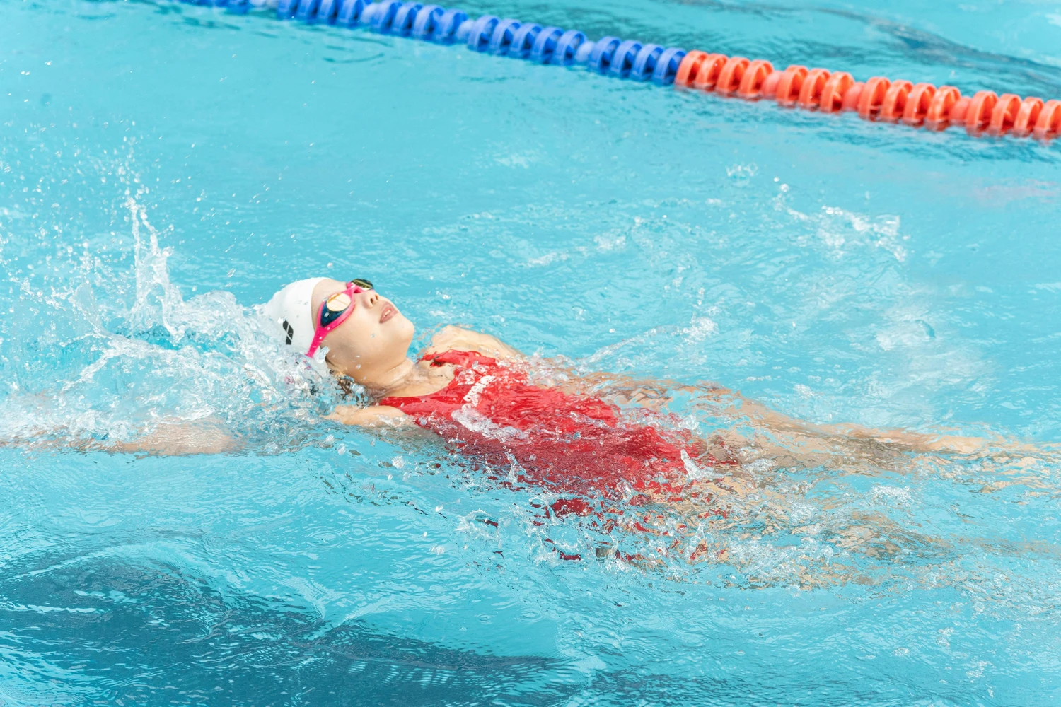 Hai Five - Schwimmschule - Eine junge Schwimmerin in rotem Badeanzug, weißer Badekappe und Schwimmbrille schwimmt Rückenschwimmen im Becken.