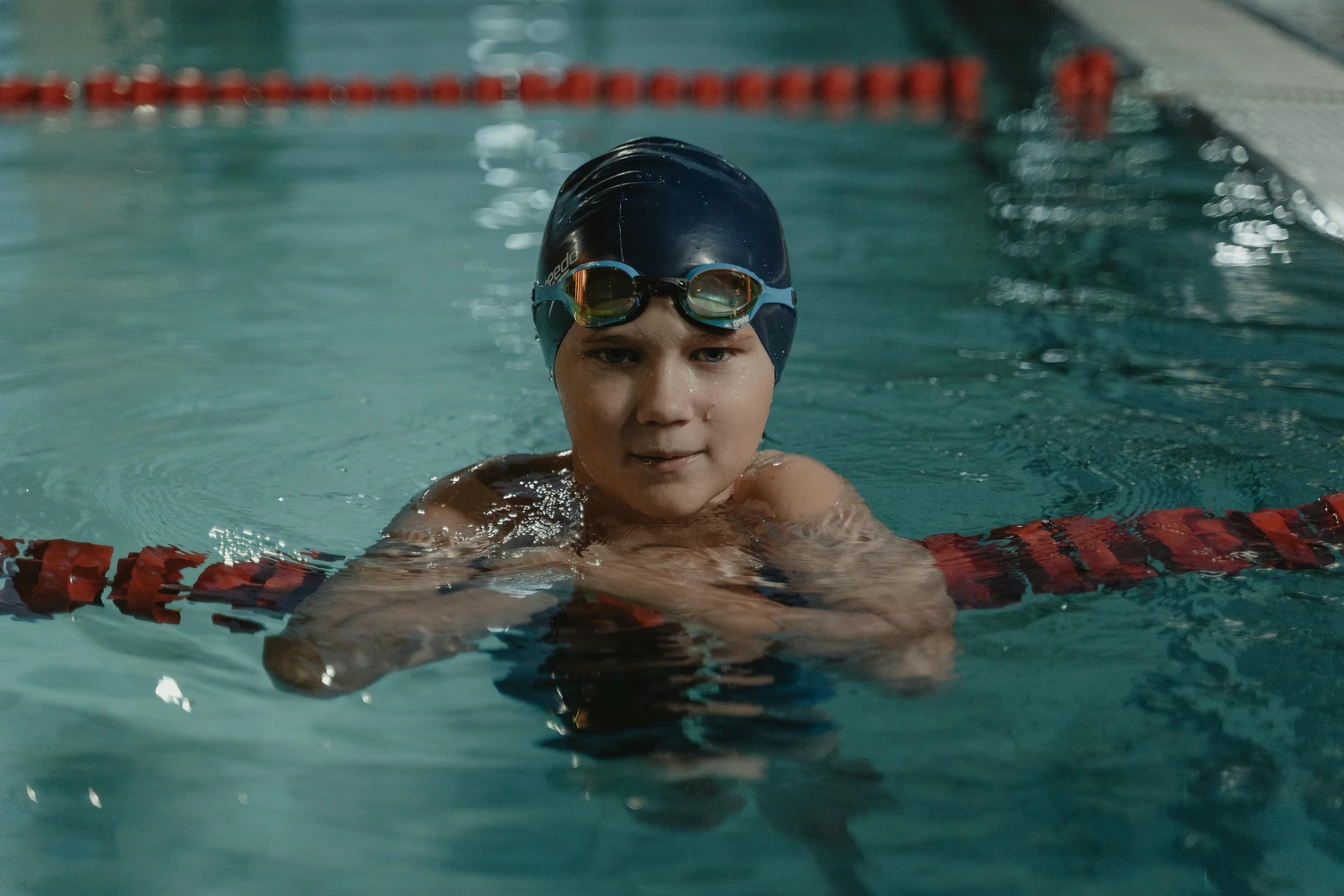 Hai Five - Schwimmschule - Ein junger Schwimmer mit Badekappe und Schwimmbrille lehnt an einer Bahnbegrenzung in einem Hallenbad.